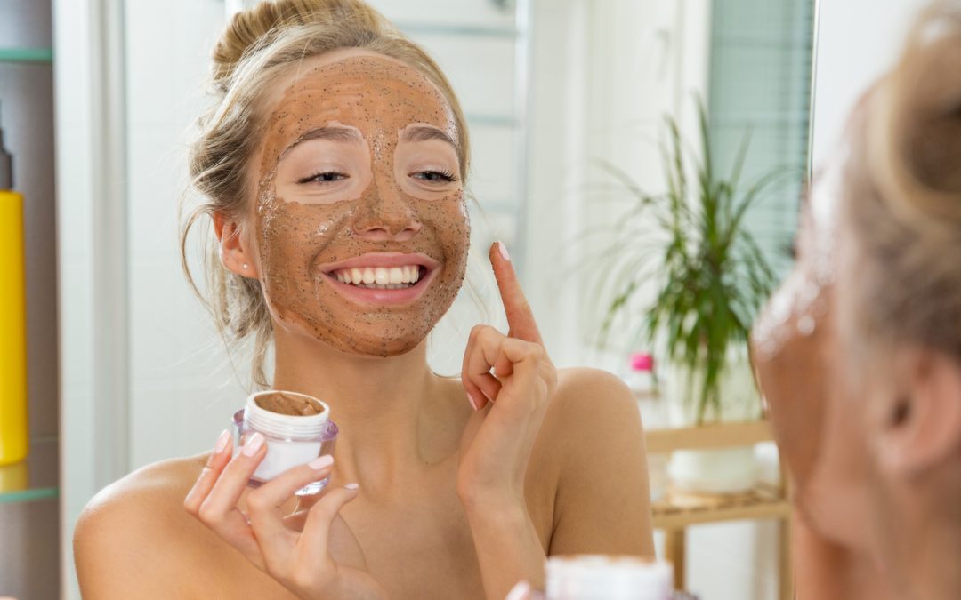 A young girl smiles at herself in the mirror as she applies a facial scrub mask at home.