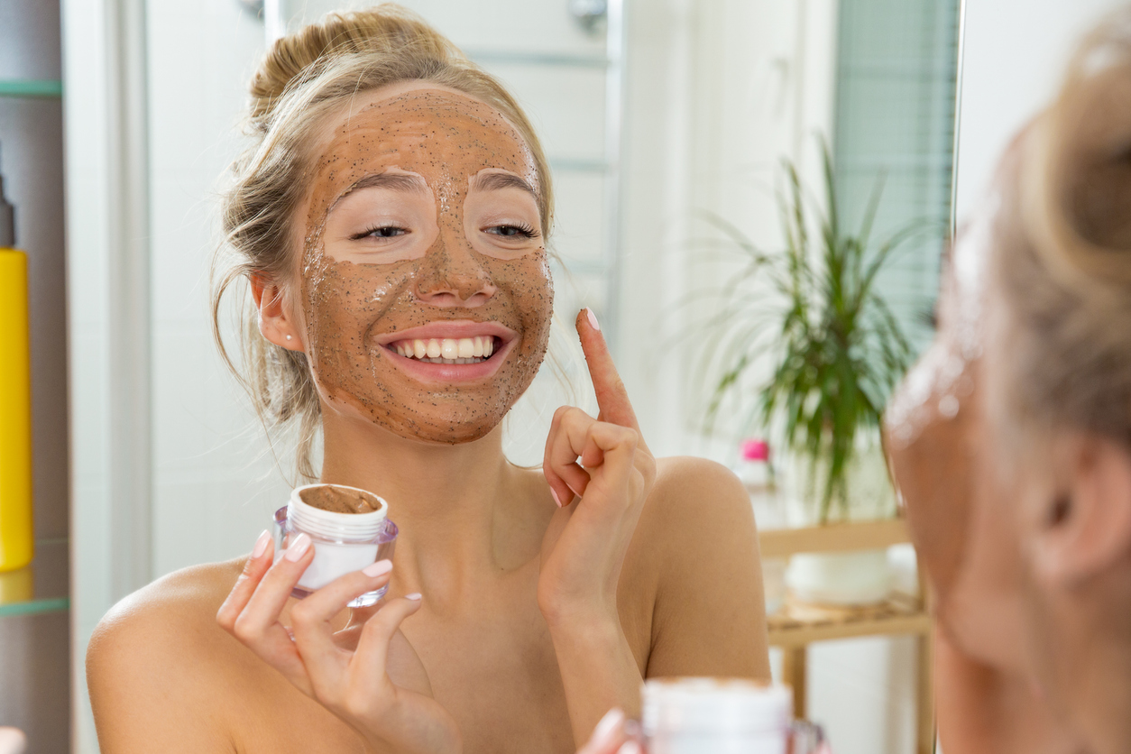 A young girl smiles at herself in the mirror as she applies a facial scrub mask at home.